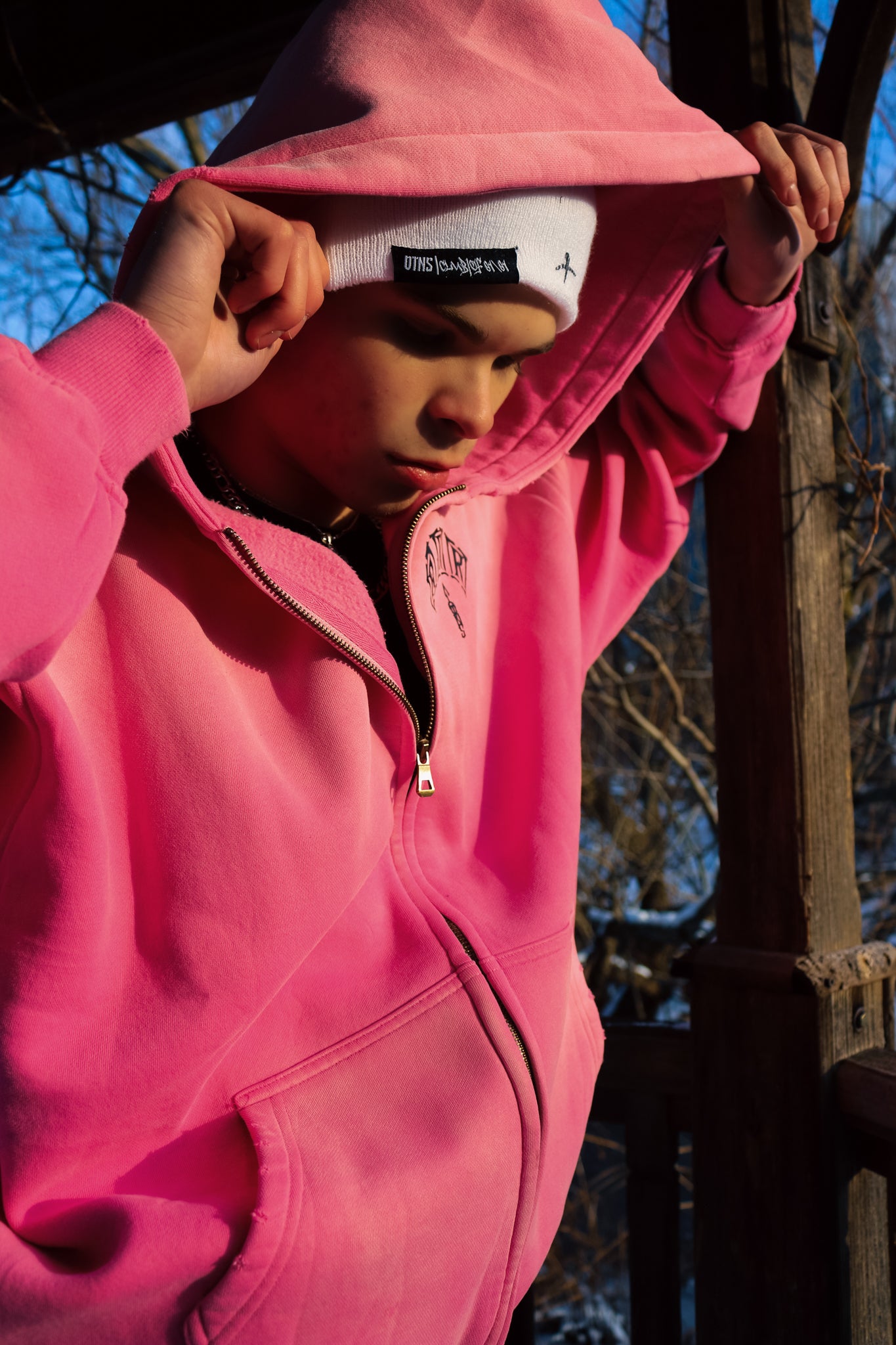 Person wearing a pink hoodie and a beanie, looking down outdoors, showcasing a relaxed streetwear style.