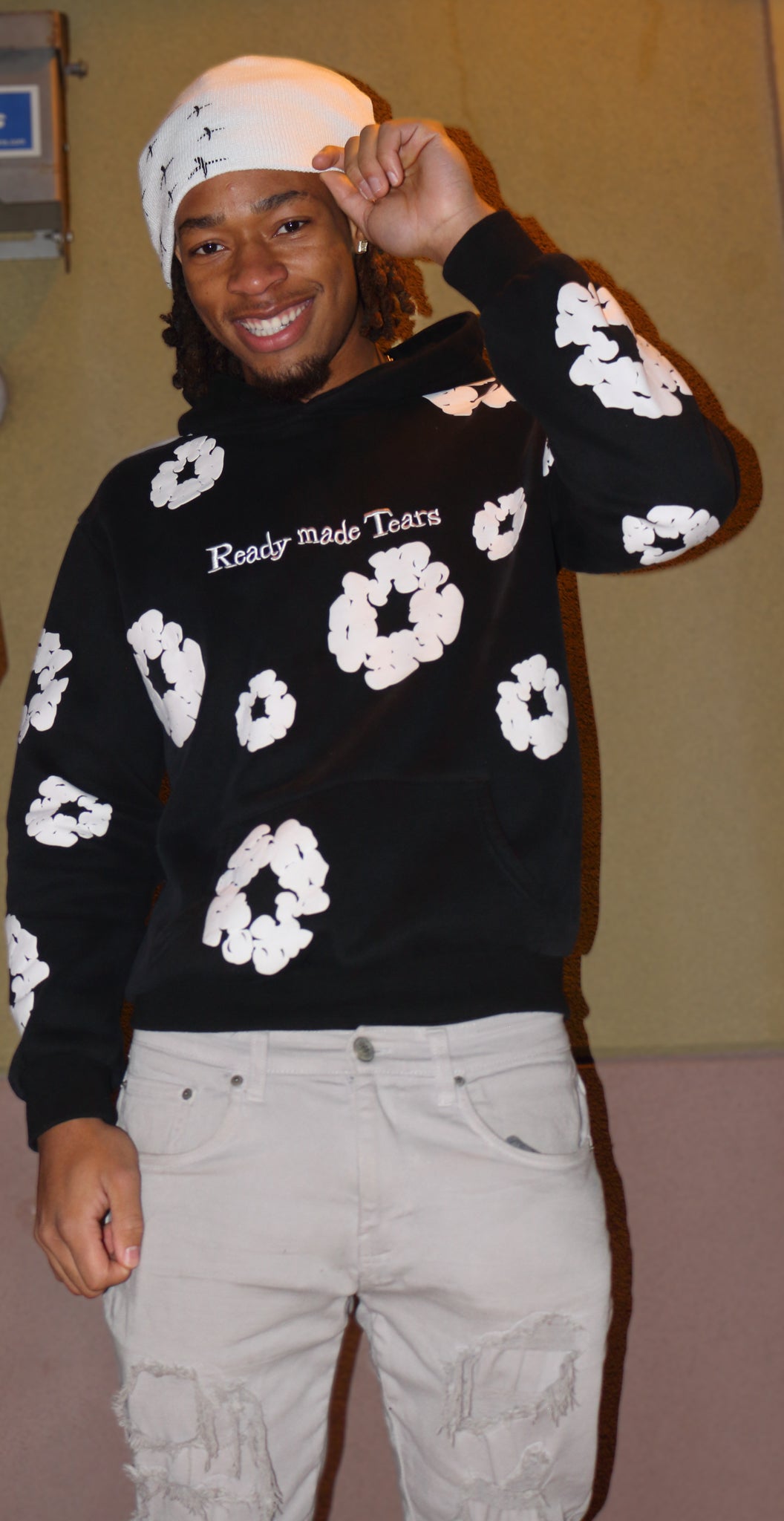 Smiling person wearing a black hoodie with floral designs and a white beanie, showcasing urban fashion style.